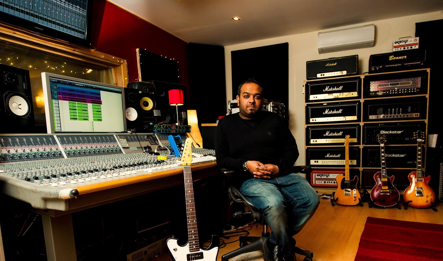 Man sitting at analogue mixing console surrounded by recording gear, guitars etc
