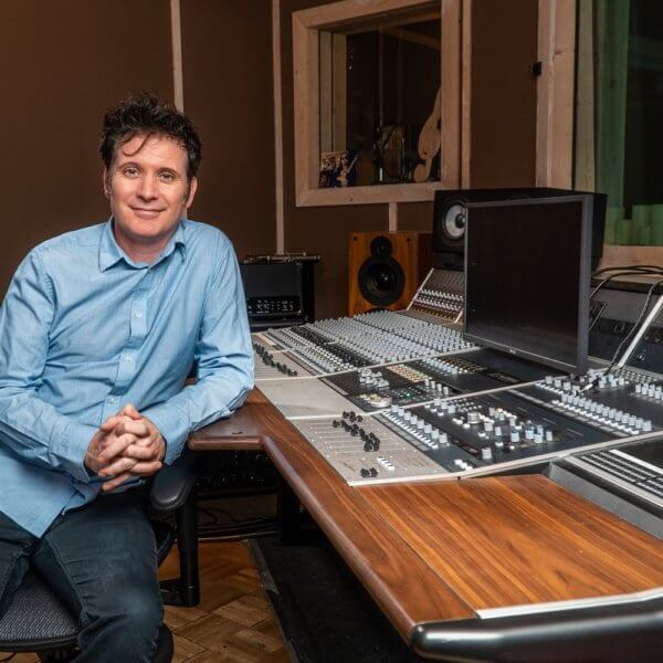 Man sits, leaning on mixing desk, looks cheerfully at camera